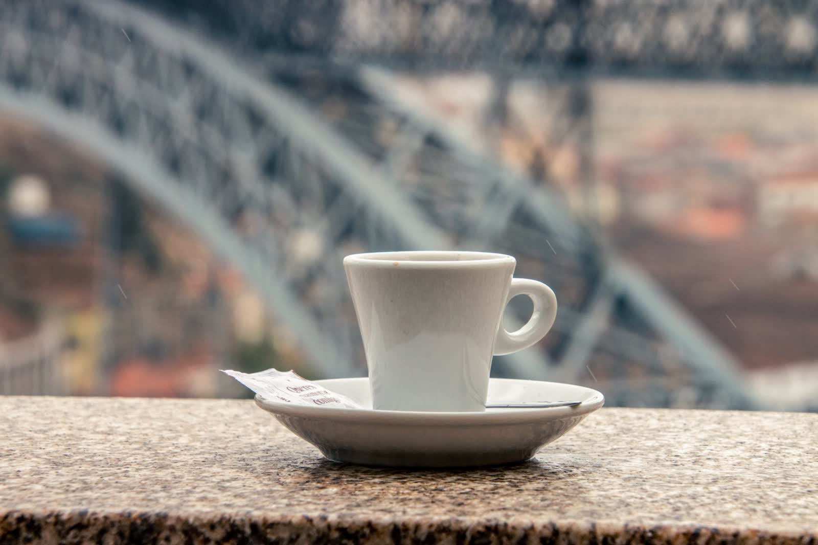 A white espresso cup on a stone ledge with a blurred iron bridge behind it