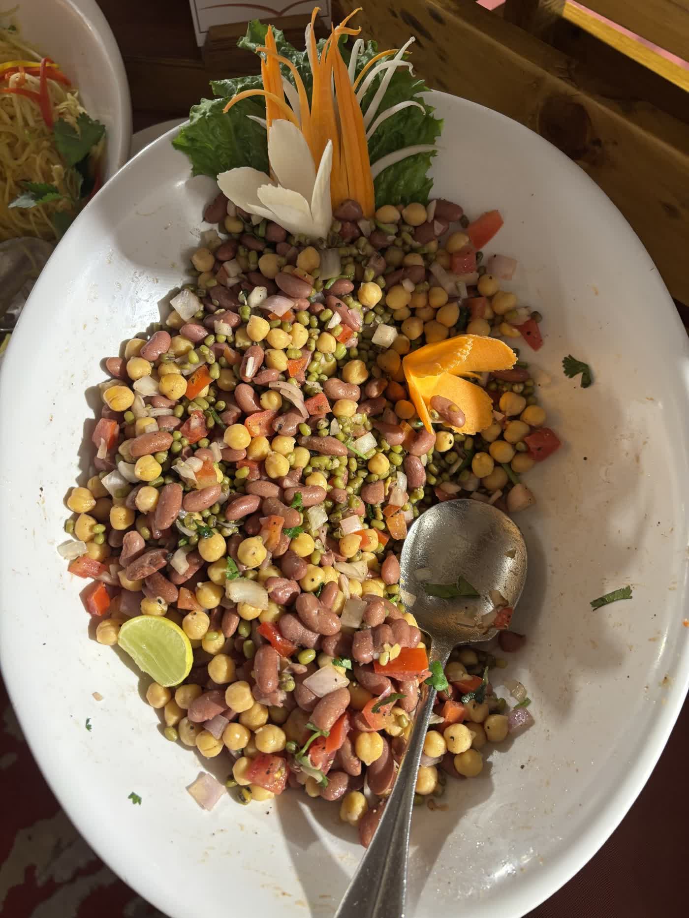 A white serving dish of mixed chickpea, kidney bean and moong salad with chopped tomato and onion, a lime wedge and carved carrot garnish