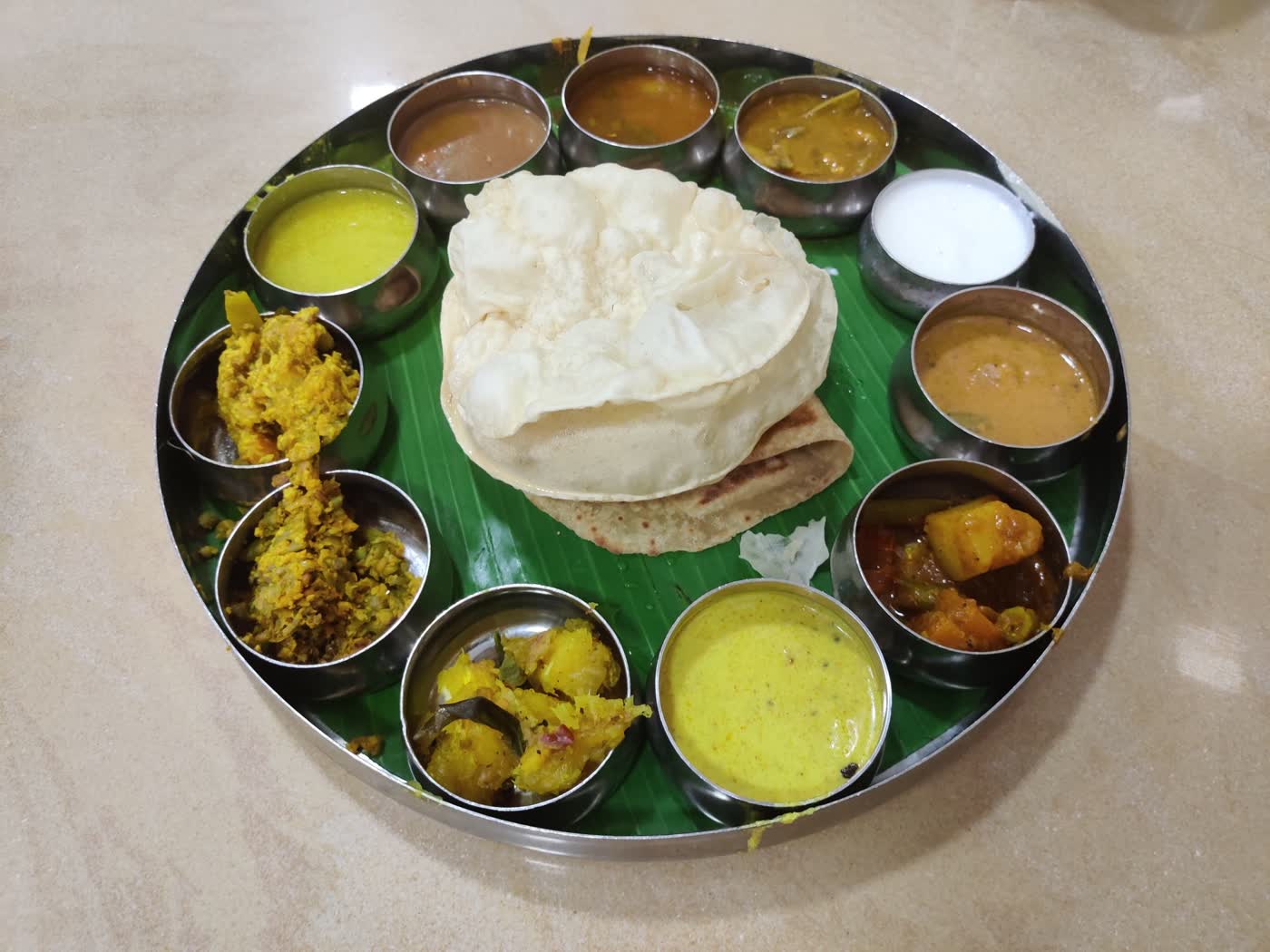 A round steel thali tray with a ring of small steel katori bowls of curries, a papad and roti in the centre