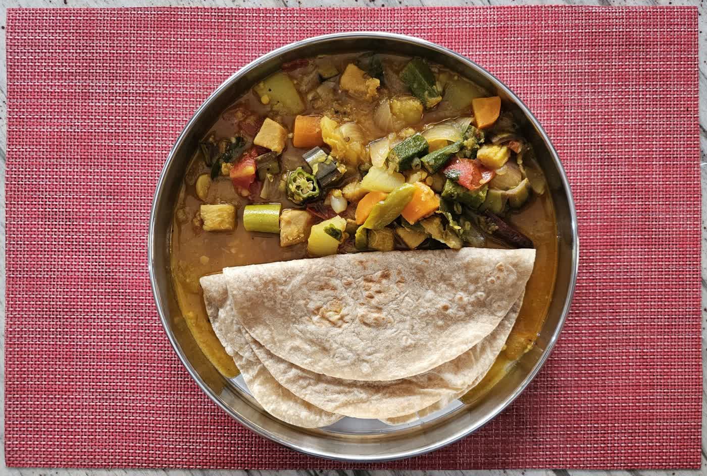 A steel plate with folded chapati and a mixed vegetable and lentil stew with okra, carrot and potato on a red placemat