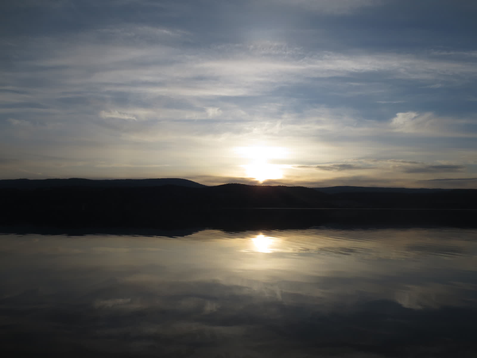 Sun setting low over a calm lake and dark hills, with the light reflected on still water