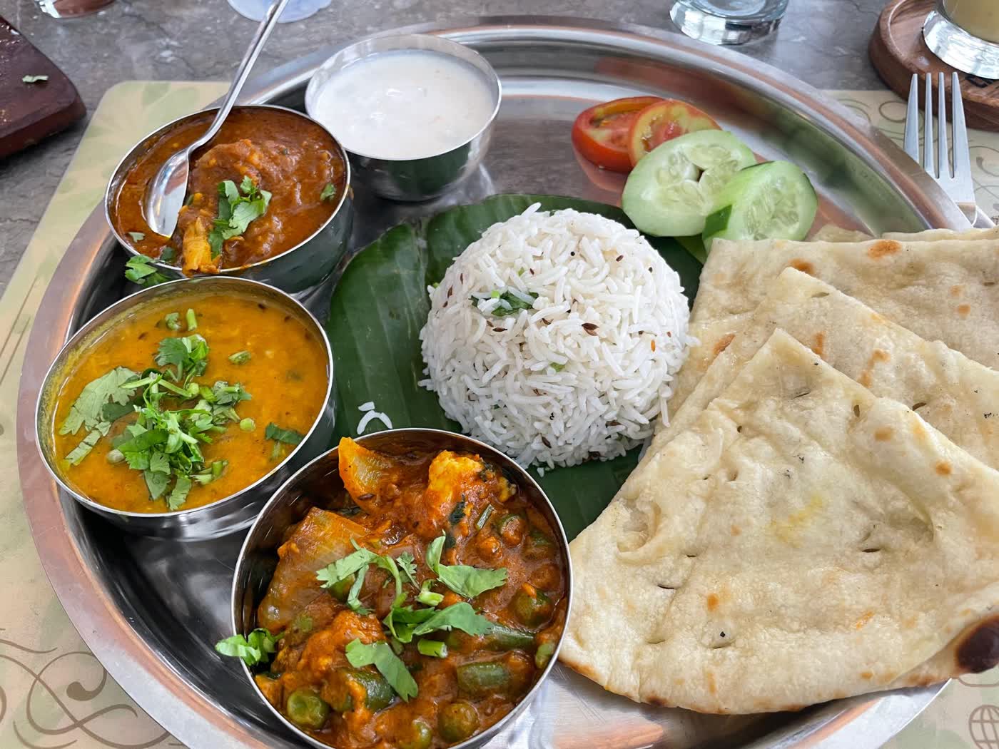 A steel thali plate with jeera rice, naan, dal, a vegetable curry, raita, tomato and cucumber