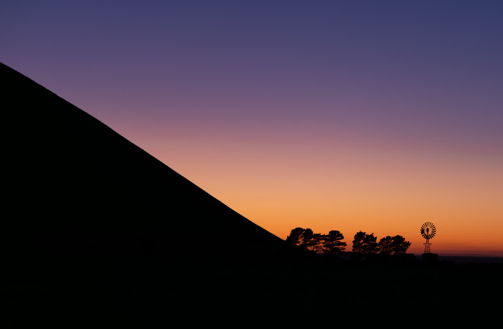 A dark hill silhouetted against an orange and purple dawn sky with a small windmill on the horizon