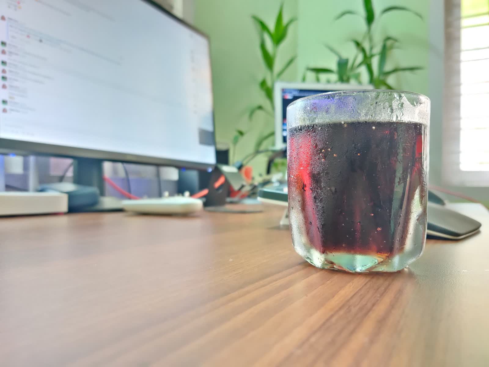 A glass of cold drink on a wooden desk in front of a monitor and green plants